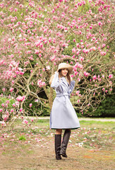 a young, beautiful woman, in a blue coat and a light hat, walks in the Park in the spring near a flowering Magnolia tree, on a Sunny day © khanfus