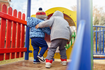 Small two caucasian boys back view in the park on the playground in the red tunnel tube wearing winter coat in autumn day having fun playing walking friendship
