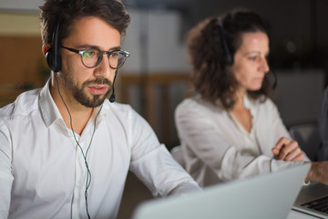 Front view of call center operator communicating with client. Confident young man in eyeglasses looking at screen of laptop and talking. Call center concept