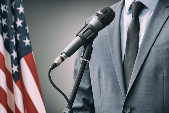 Man in suit standing close to microphone with USA flag on background