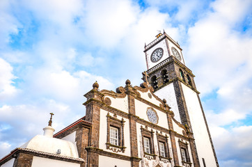 The outdoor facade of St. Sebastian Church, Igreja Matriz de Sao Sebastiao, in Ponta Delgada, Azores, Portugal. White clock tower from below with blue sky and clouds above. Sunset light, golden hour