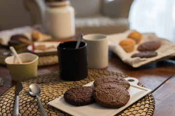 A rustic food set with homemade chocolate biscuits on a tray next to some mugs, cookies and muffins in a defocused background
