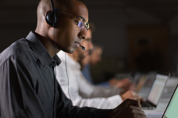 Focused call center operator typing on laptop. African American young man in eyeglasses looking at...