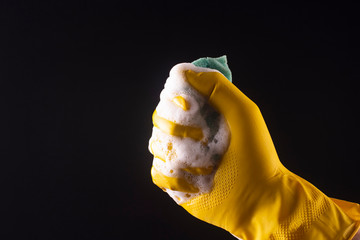 A hand in a yellow latex glove squeezes foam from a washcloth on a black background ..