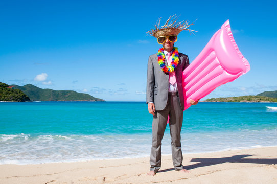 Smiling businessman wearing straw hat and oversized sunglasses standing with a pink lilo on a tropical beach