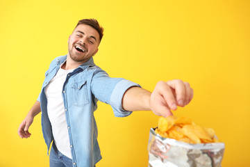 Handsome young man eating tasty potato chips on color background