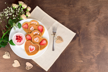 Homemade heart shaped pancakes with strawberry cream and berry slices on a white plate served on a wooden table. Rustic style breakfast or brunch for Valentine's Day. Top view, copyspace