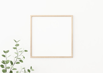 Interior poster mockup with square wooden frame on empty white wall decorated with plant branch with green leaves. 3D rendering, illustration.