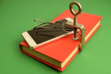 la lettura dei libri è stata sostituita dalla connessione dello smartphone ad Internet : libro chiuso da una morsa, uno smartphone  e un paio di occhiali