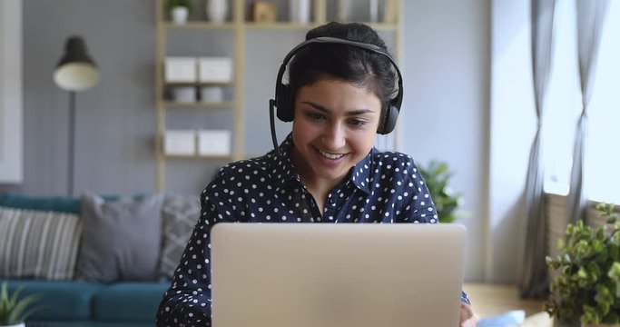 Smiling indian woman wear wireless headset video calling on laptop