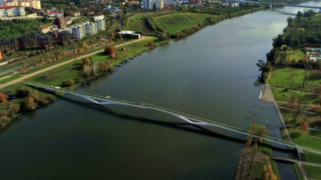 Drone footage of Ponte Pedonal Pedro e Ines bridge and Mondego river in Coimbra, Portugal surrounded by green grass river banks and city buildings.