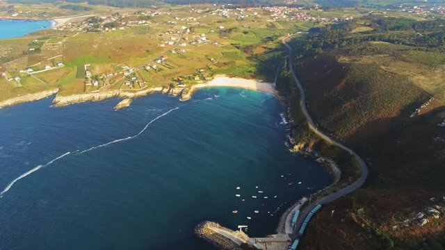 Drone footage of Playa de Barizo in Malpica, Spain with boat station at the base of green hill with deep blue turquoise water and warm sunlight