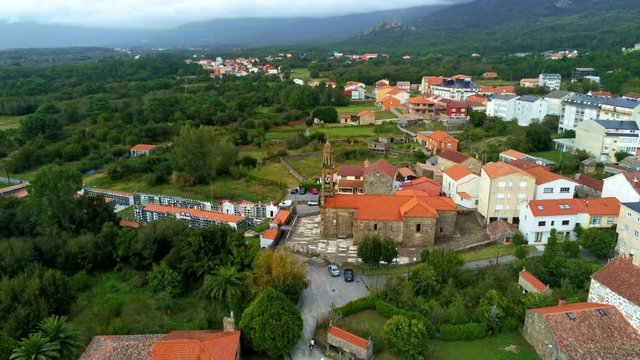 Drone footage of Iglesia de Santa Comba de Carnota in A Coruna, Spain on a partly cloudy day surrounded by green trees and birds flying in and out of frame