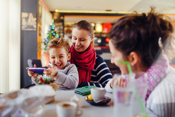 Small little boy caucasian child son sitting in the lap of his mother or aunt while female friend is looking at them teasing smiling holding smart mobile phone playing games or making a video call