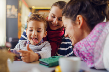 Fototapeta premium Small little boy caucasian child son sitting in the lap of his mother or aunt woman while female friend is looking to the smart mobile phone playing games or making a video call