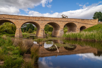 Fototapeta na wymiar Richmond, Tasmania, Australia - December 13, 2009: Aong water line closeup shot of brown stone historic bridge over coal river reflected in water with reed and green lawn on side. Blue cloudscape.