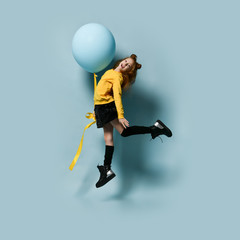 Fototapeta na wymiar Teenager in yellow sweatshirt, black skirt, knee-highs, boots. She smiling, holding balloon, jumping up against blue background.