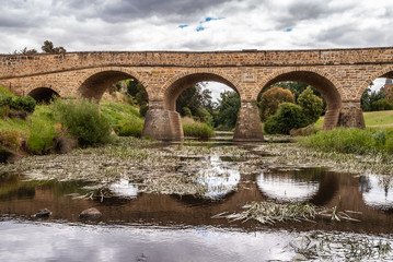 Fototapeta na wymiar Richmond, Tasmania, Australia - December 13, 2009: Aong water line shot of brown stone historic bridge over coal river reflected in water with reed and green lawn on side. Dark cloudscape.