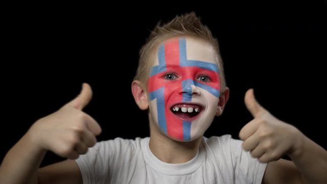Happy boy rejoices victory of his favorite team of Faroe Islands. A child with a face painted in national colors. Portrait of a happy young fan. Joyful emotions and gestures. Victory. Triumph.