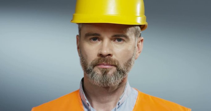 Caucasian handsome gray-haired builder with a beard and in yellow helmet turning face to the camera and looking straight to the camera. Close up.