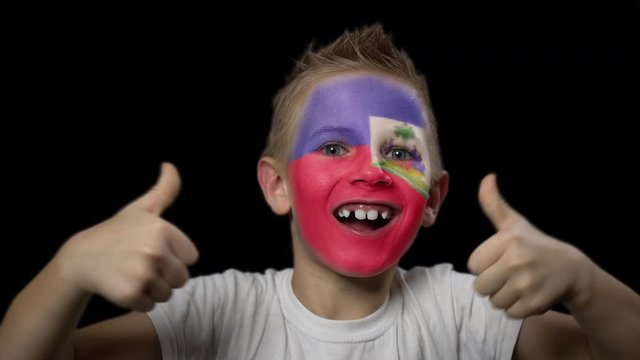Happy boy rejoices victory of his favorite team of Haiti. A child with a face painted in national colors. Portrait of a happy young fan. Joyful emotions and gestures. Victory. Triumph.
