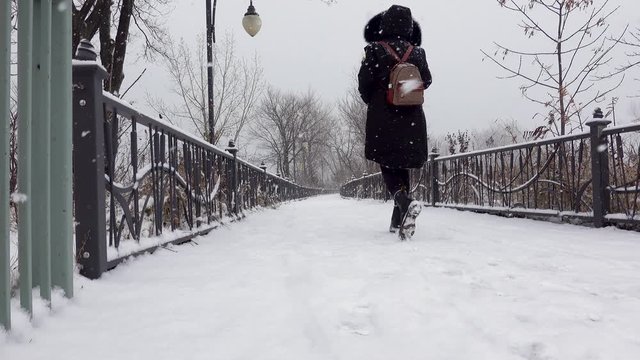 a heavy snow fall on a pedestrian bridge in an urban park and a woman wearing black coat and a brown backpack walking into the scene and she starts taking pictures in the middle of the bridge