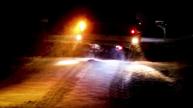 Snow plow truck at night spreading salt during snowstorm going uphill residential neighborhood fades black