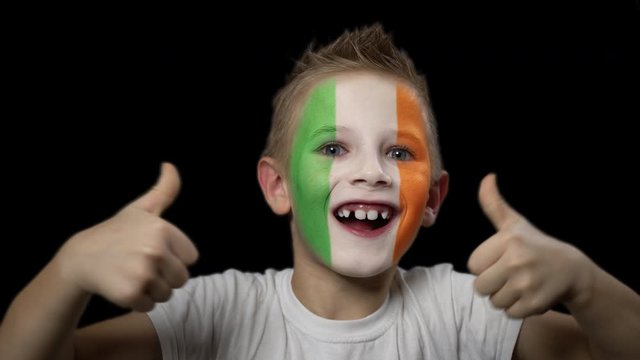 Happy boy rejoices victory of his favorite team of Ireland. A child with a face painted in national colors. Portrait of a happy young fan. Joyful emotions and gestures. Victory. Triumph.