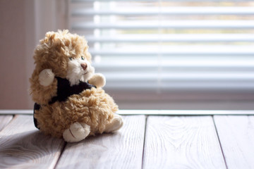 A brown teddy bear in a black sweater is sitting on a wooden table by the window.