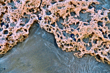 Ocean bed rock uncovered by an ebbing tide, pink and coral porous rock and wet sand marked with...