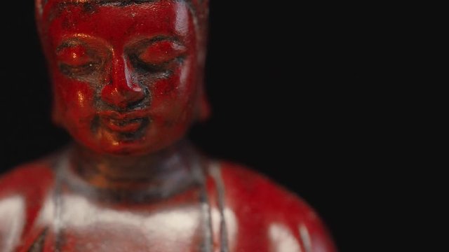 Dolly camera move along sitting in meditation statue of Buddha. Figure is lit by a candle flame on dark background. Panning shot from left to right, slowly sliding, close-up shot.