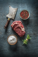 Raw beef steaks with meat cleaver and salt pepper on dark concrete background