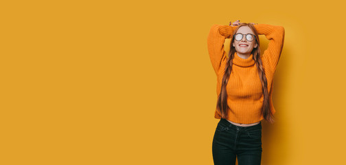 Beautiful young woman with red hair and freckles dressed in yellow looking at camera smiling while leaning on a yellow studio wall.