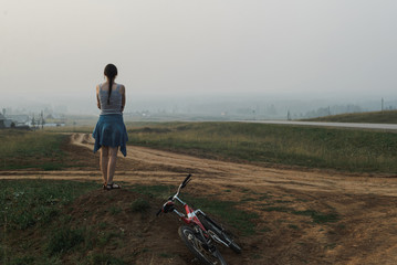 white caucasian young woman standing in village hill road with bike, looking at open space, view from back in full body size, horizontal lifestyles stock photo image with copy space for text