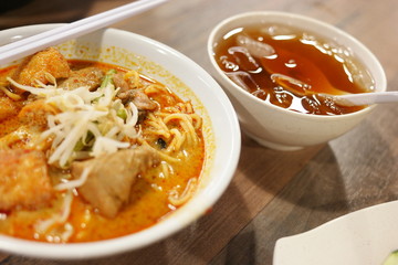 Delicious bowl of curry laksa with noodles, tofu puffs, and chicken, served with a side drink.
