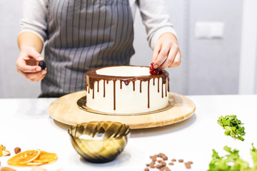 Confectioner decorates with berries a biscuit cake with white cream and chocolate. Cake stands on a wooden stand on a white table. The concept of homemade pastry, cooking cakes.