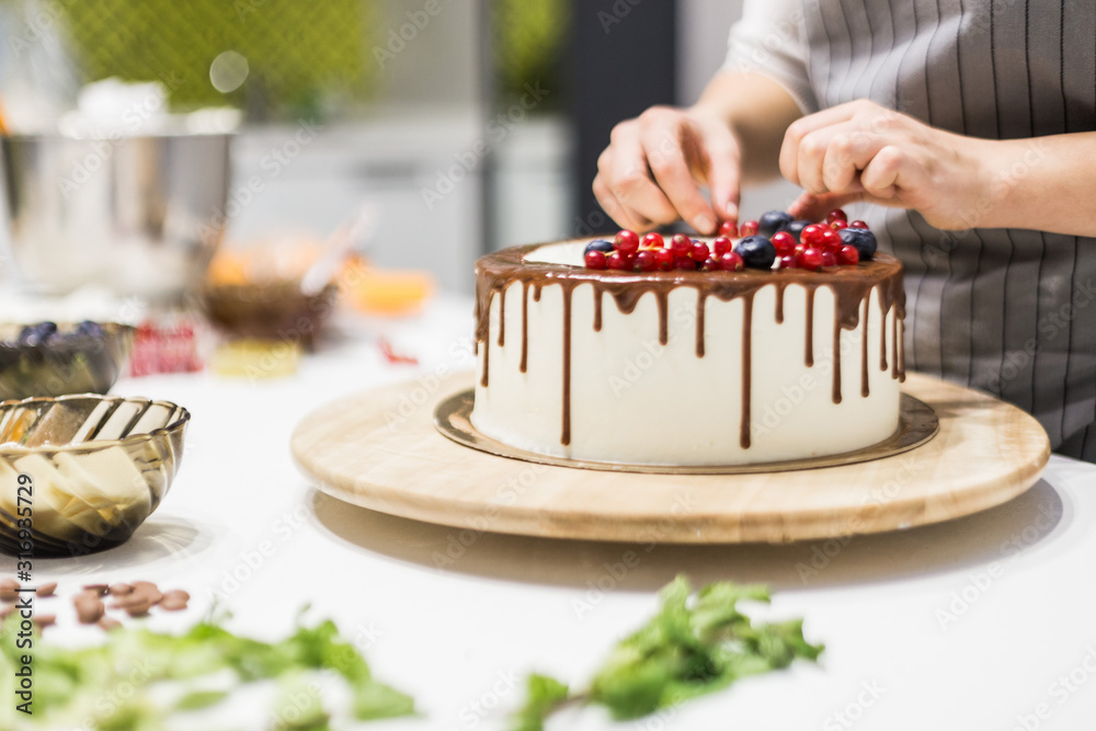 Wall mural confectioner decorates with berries a biscuit cake with white cream and chocolate. cake stands on a 