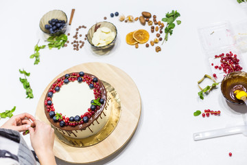 Confectioner decorates with berries a biscuit cake with white cream and chocolate. Cake stands on a wooden stand on a white table. The concept of homemade pastry, cooking cakes.