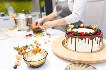 Confectioner decorates with berries a biscuit cake with white cream and chocolate. Cake stands on a wooden stand on a white table. The concept of homemade pastry, cooking cakes.