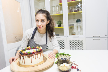 A confectioner stands next to a cooked biscuit cake with white cream, decorated with chocolate and berries. Cake stands on a wooden stand on a white table