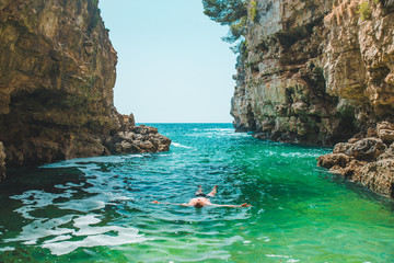 young man swimming on back in sea at bay between cliffs