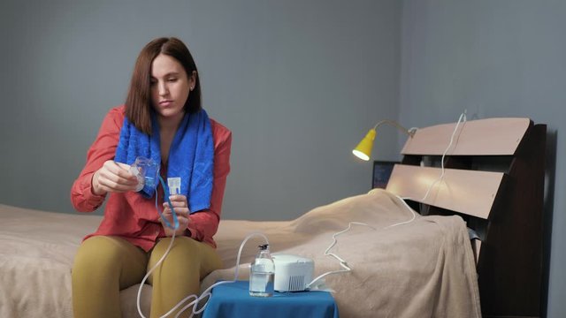 Girl screws inhalation mask to nebulizer. Young woman dressed in red shirt and blue towel sits alone in room and fastens mask to inhaler. Slow motion and general plan