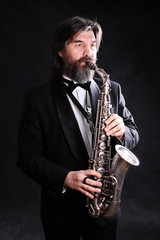 Fototapeta premium A male artist musician in a classic black suit, tailcoat, statuesque in a bow tie with a beard plays music on a gold saxophone.black background
