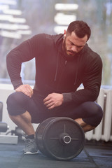 Fototapeta na wymiar tired strong young caucasian man with beard in black sportswear sit with iron barbell relaxing after heavy intensive workout training in gym