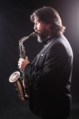 Fototapeta na wymiar A male artist musician in a classic black suit, tailcoat, statuesque in a bow tie with a beard plays music on a gold saxophone.Playing an instrument against a white light source.black background