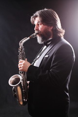 Obraz na płótnie Canvas A male artist musician in a classic black suit, tailcoat, statuesque in a bow tie with a beard plays music on a gold saxophone.Playing an instrument against a white light source.black background