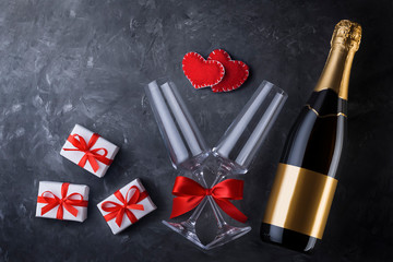 Valentine's day greeting card template from champagne bottle, two luxury glasses with a red satin ribbon bow, two handmade hearts and three gift boxes with copy space on black texture background.