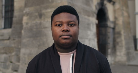 Obraz premium Front view of confident man standing at old city street. African guy looking to camera with serious face and blinking. Portraits. Concept of people and emotions. Outdoors.