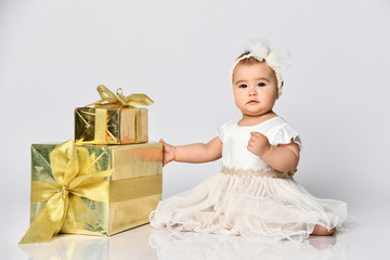 Fototapeta premium Baby girl in dress and headband, barefoot. She holding golden gift box, sitting on floor isolated on white. Close up, copy space