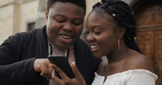 Close up of cheerful couple looking at smartphone screen and laughing. Young african couple standing outdoors using mobilephone, smiling, talking. Concept of people and coomunication.